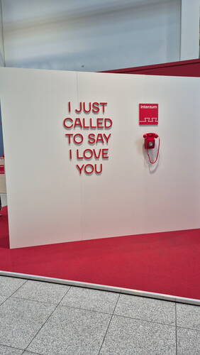 Rote Telefoninstallation auf einer Messewand mit dem Schriftzug „I JUST CALLED TO SAY I LOVE YOU“ über rotem Teppichboden