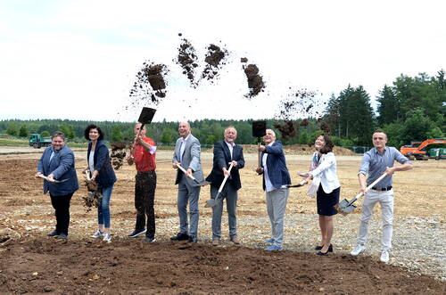 Gruppe von acht Personen in business- und Freizeitkleidung wirft beim symbolischen ersten Spatenstich auf einer Baustelle gleichzeitig Erde in die Luft, im Hintergrund Wald und Baumaschinen