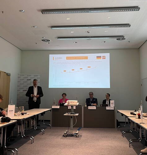 Geschäftspräsentation in einem Konferenzraum, bei der ein Referent vor einer Leinwand mit Wachstumsdiagramm zur Küchenbranche spricht, während drei weitere Personen am Podium sitzen und zuhören