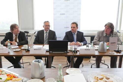 Vier Männer im Anzug sitzen mit Namensschildern an einem Konferenztisch, diskutieren vor Laptops und Mikrofonen in einem hellen Besprechungsraum mit Getränken und Snacks.