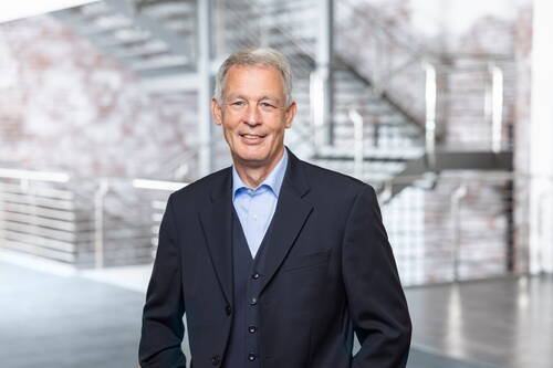 Älterer Mann im dunkelblauen Anzug lächelt freundlich in moderner Bürohalle mit metallener Treppe im Hintergrund