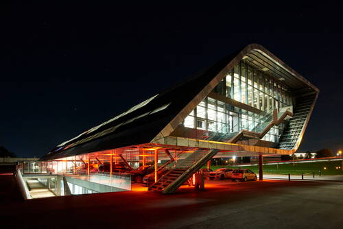 Futuristisches, verglastes Bürogebäude mit schrägem Dach und beleuchtetem Parkdeck in warmem Rotlicht bei Nacht