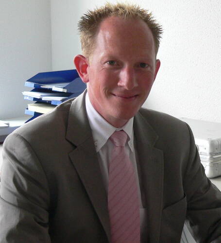 Junger Geschäftsmann mit kurzem, blondem Haar im grauen Anzug und rosa Krawatte sitzt lächelnd an einem Schreibtisch vor einem Stapel Aktenordner.