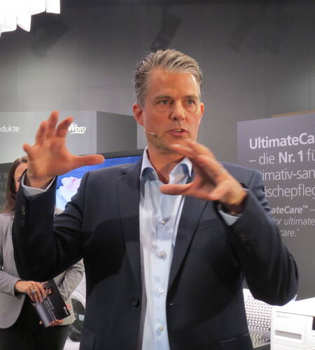 Mittelalter Mann im Anzug erklärt mit ausladenden Handbewegungen ein Produkt auf einer Messepräsentation vor einem Messestand mit technischen Geräten und Werbetafeln