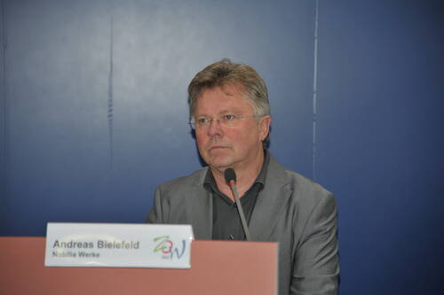 Mittelalterlicher Mann im grauen Anzug sitzt ernst hinter einem Rednerpult mit Namensschild und Mikrofon vor einer blauen Wand