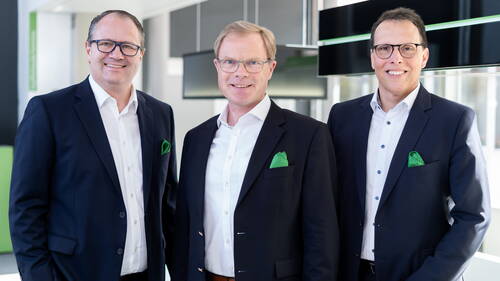 Drei lächelnde Geschäftsleute in dunklen Anzügen mit grünen Einstecktüchern stehen nebeneinander in einem modernen, hellen Büro.