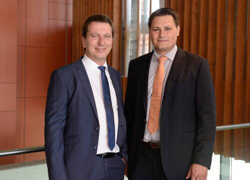 Zwei Geschäftsmänner im Anzug stehen nebeneinander in einem modernen Bürogebäude mit Holzvertäfelung und Glasgeländer und blicken lächelnd in die Kamera.