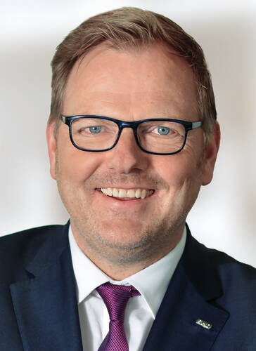 Lächelnder Mann mittleren Alters mit Brille im blauen Anzug und lila Krawatte vor hellem Hintergrund