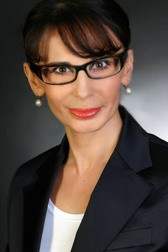 Elegant gekleidete Frau mit dunklem Haar, Brille und Perlenohrringen, die vor dunklem Hintergrund direkt in die Kamera blickt