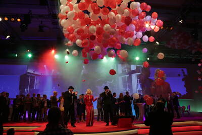 Bunte Bühnenveranstaltung mit Sängerin im roten Glitzerkleid, umgeben von festlich gekleideten Menschen, während rote und weiße Luftballons von der Decke herabfallen
