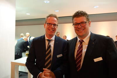 Zwei lächelnde Männer in Anzügen und Namensschildern stehen bei einer geschäftlichen Abendveranstaltung in einem hell beleuchteten Konferenzraum, während im Hintergrund weitere Gäste miteinander sprechen.
