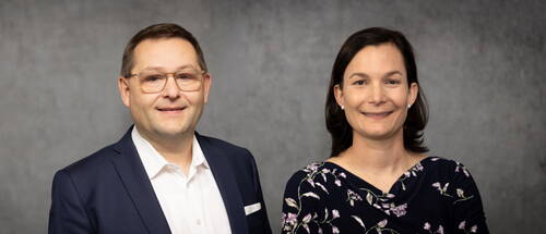 Zwei lächelnde Personen posieren vor grauem Hintergrund, der Mann links im dunkelblauen Anzug mit weißem Hemd, die Frau rechts im dunkelblauen Kleid mit floralem Muster.
