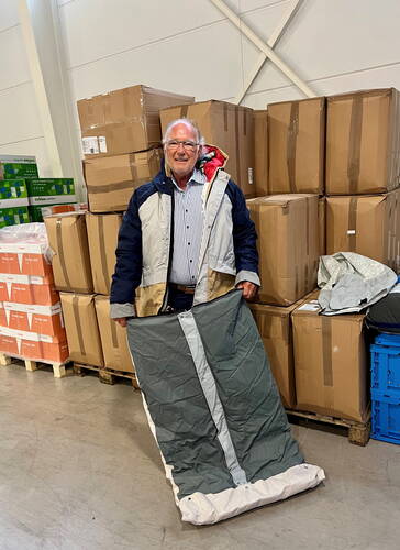 Älterer Mann im Lager steht vor gestapelten Kartons und präsentiert einen ausgebreiteten, grauen Schlafsack auf dem Boden