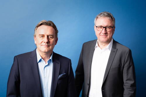 Zwei mittelalte Männer in Business-Anzügen stehen lächelnd nebeneinander vor einem einfarbig blauen Hintergrund.