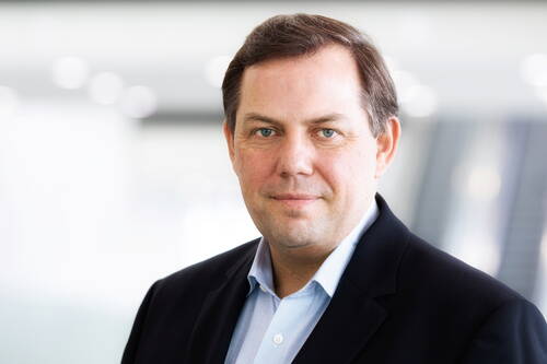 Mittelalterlicher Mann im Business-Anzug blickt ruhig in die Kamera, vor einem hellen, leicht verschwommenen Bürohintergrund
