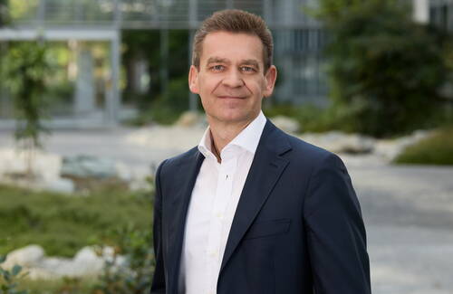 Mittelalterlicher Mann im dunklen Anzug und weißem Hemd lächelt selbstbewusst im modernen, begrünten Firmenhof