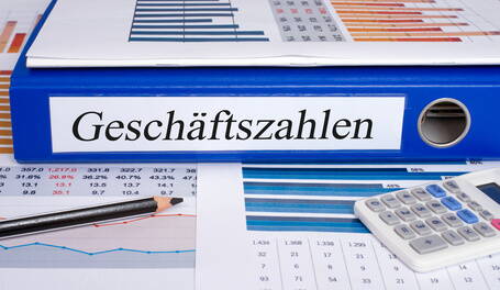 Blauer Ordner mit der Aufschrift „Geschäftszahlen“ liegt auf ausgedruckten Diagrammen und Tabellen neben Taschenrechner und Bleistift