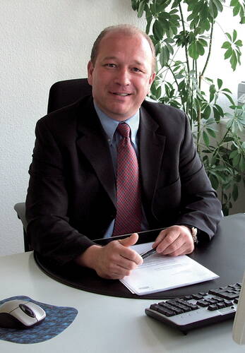 Geschäftsmann im Anzug lächelt an seinem Schreibtisch, hält einen Stift über Unterlagen vor Computer und Zimmerpflanze