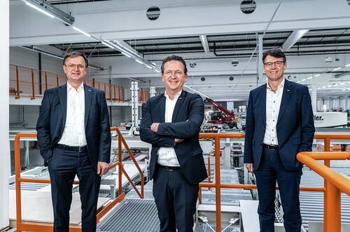 Drei Geschäftsleute im Anzug stehen lächelnd in einer modernen Industriehalle auf einer erhöhten Plattform mit orangefarbenem Geländer