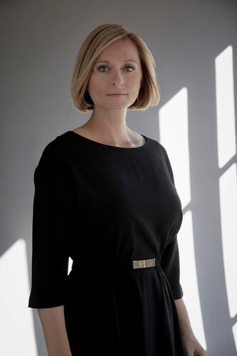 Frau mit blondem Bob und schwarzem Kleid steht ernst blickend im weichen Seitenlicht vor einer grauen Wand mit hellen Fensterschatten