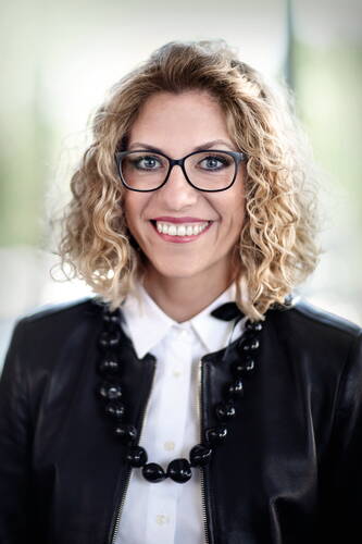 Lächelnde Frau mit lockigen blonden Haaren, schwarzer Brille, weißer Bluse und schwarzer Lederjacke vor unscharfem, hellgrünem Hintergrund