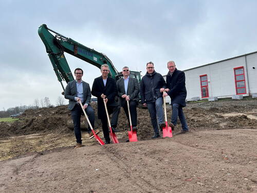 Fünf Männer in Anzügen und Mänteln führen mit roten Spaten einen symbolischen ersten Spatenstich vor einem Bagger auf einer Baustelle neben einer grauen Industriehalle aus.