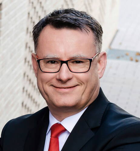 Lächelnder Mann mit Brille im dunklen Anzug und roter Krawatte vor unscharfem städtischem Hintergrund