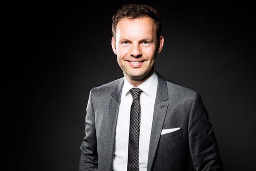 Lächelnder Mann im grauen Businessanzug mit Krawatte vor dunklem Hintergrund in professioneller Porträtaufnahme