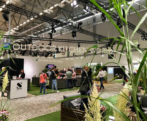 Besucher unterhalten sich und testen Grillstationen am Messestand „Outdoor Kitchen World“ in einer großen, modern beleuchteten Ausstellungshalle mit viel Grün und Dekopflanzen.