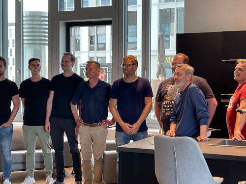 Gruppe lächelnder Männer steht locker in einer modernen Loftküche vor großen Fenstern mit Stadtblick