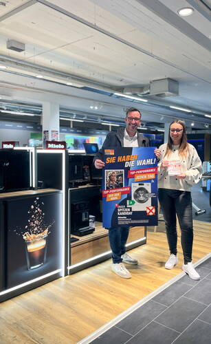 Zwei Mitarbeitende in einem Elektromarkt präsentieren gemeinsam ein großes, buntes Werbeplakat für Miele-Haushaltsgeräte neben einer modernen Kaffeemaschinen-Ausstellung.