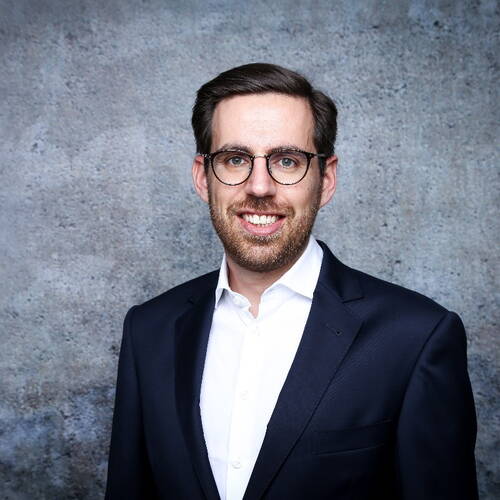 Junger Mann mit Brille und dunklem Anzug lächelt vor grauer Betonwand in einem professionellen Porträt.