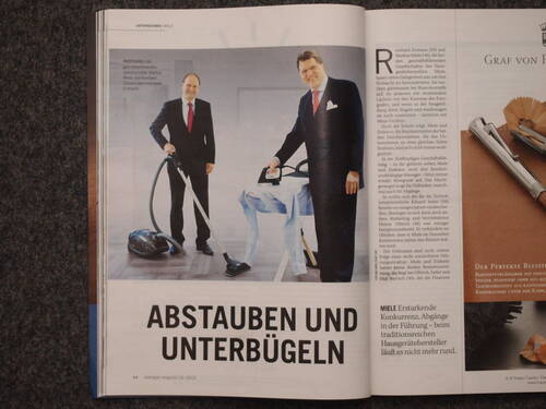 Zwei Geschäftsleute im Anzug präsentieren in einem Magazinartikel staubsaugende und bügelnde Hausgeräte neben der Überschrift „Abstauben und Unterbügeln“