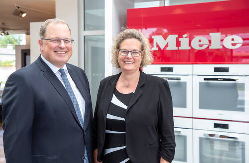 Zwei lächelnde Geschäftspersonen im Anzug stehen in einer modernen Ausstellung vor weißen Einbauöfen mit rotem Miele-Logo im Hintergrund.