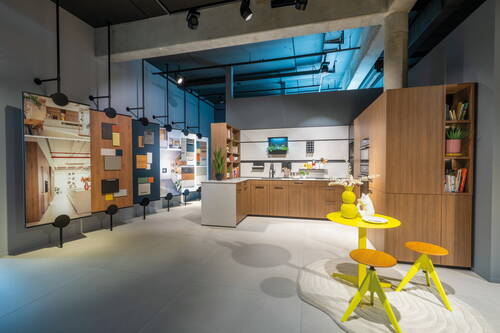 Modernes Küchenstudio mit holzvertäfelter Einbauküche, hellen Arbeitsplatten, Musterwänden für Fronten und Arbeitsflächen sowie einem kleinen gelben Bistrotisch mit zwei Hockern im Vordergrund
