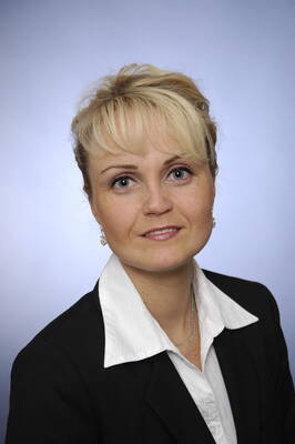 Blonde Frau im Business-Anzug mit weißer Bluse blickt freundlich in die Kamera vor neutral blauem Hintergrund