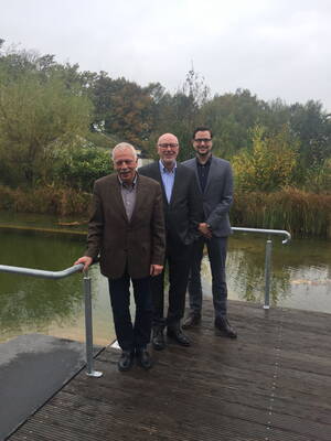 Drei Männer in Anzügen stehen lächelnd auf einem Holzsteg vor einem Teich mit herbstlich gefärbten Bäumen im Hintergrund