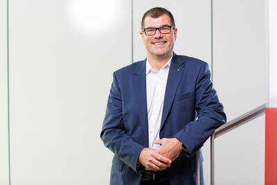 Lächelnder Mann im blauen Business-Anzug mit Brille steht an einem Treppengeländer in einem hellen, modernen Büro.