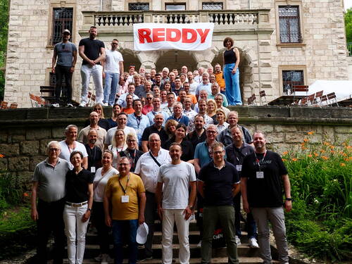 Große Gruppe von Mitarbeitenden posiert lächelnd auf einer Steintreppe vor einem historischen Gebäude, über dem ein Banner mit der Aufschrift „REDDY“ hängt.