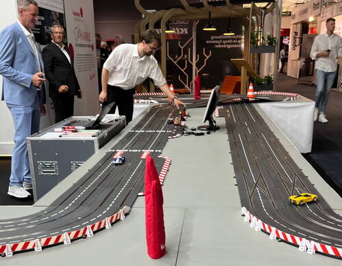Männer in Businesskleidung steuern auf einer Messe eine große Carrera-Rennbahn mit mehreren bunten Modellautos, während Besucher im Hintergrund den Stand beobachten.