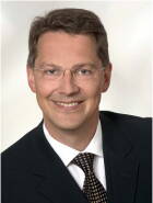 Lächelnder Mann im Anzug mit Krawatte und Brille vor neutral hellem Hintergrund