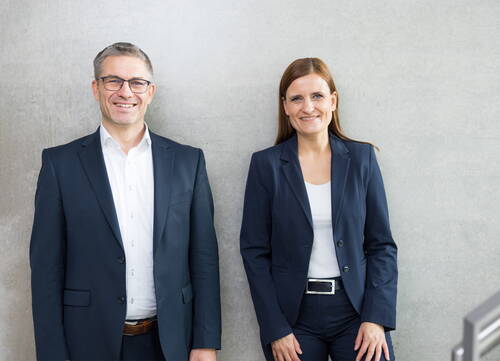 Zwei lächelnde Geschäftsleute im dunkelblauen Anzug stehen nebeneinander vor einer hellgrauen Betonwand und blicken direkt in die Kamera.