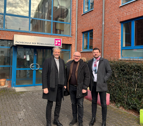 Drei lächelnde Männer in Winterkleidung stehen vor dem Eingang der „Fachschule des Möbelhandels“ an einem roten Backsteingebäude mit blauen Fensterrahmen.