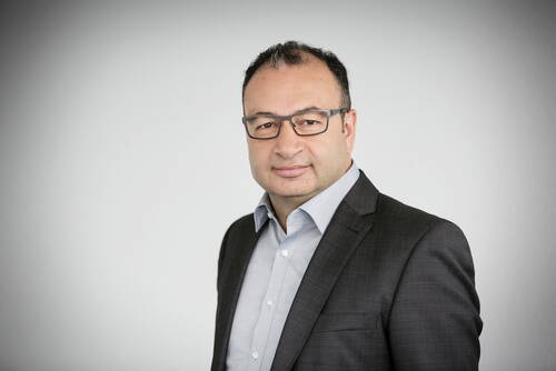Mittelalterlicher Mann mit Brille in dunklem Anzug und hellblauem Hemd blickt ruhig in die Kamera vor neutralem, hellgrauem Hintergrund