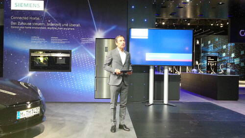 Geschäftsmann in grauem Anzug präsentiert auf einer Siemens-Messebühne ein vernetztes Zuhause mit großem Display und Smart-Home-Geräten im Hintergrund