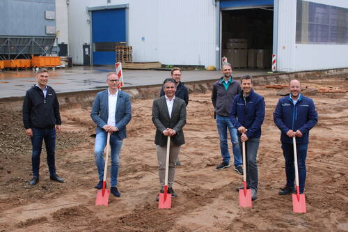 Sieben Männer stehen auf einer Baustelle vor einer Industriehalle, vier von ihnen halten rote Spaten für den offiziellen Spatenstich einer neuen Firmenerweiterung.
