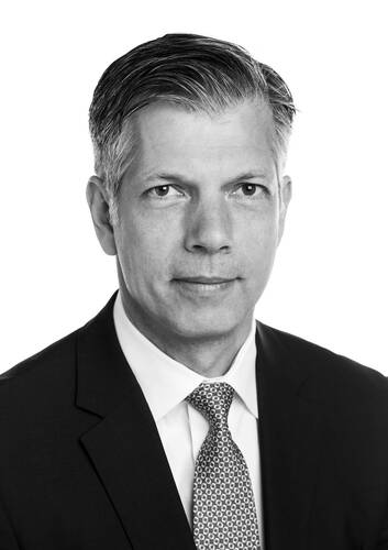 Porträt eines mittelalten Mannes im Anzug mit Krawatte, der ernst in die Kamera blickt, vor neutral weißem Hintergrund in Schwarz-Weiß-Aufnahme.