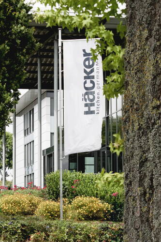 Weiße Häcker-Fahne vor dem modernen Firmengebäude, umgeben von gepflegten Grünanlagen und blühenden Sträuchern