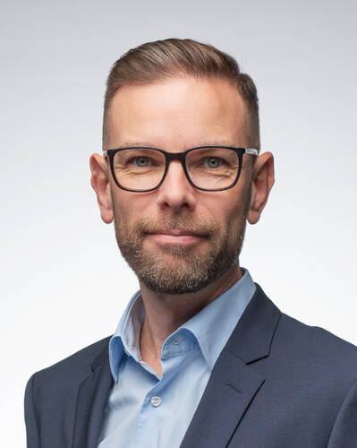 Mittelalterlicher Mann mit Kurzhaarfrisur, Vollbart und Brille in blauem Hemd und dunkelblauem Sakko blickt freundlich in die Kamera vor neutral hellem Hintergrund.