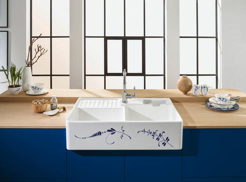 Helle Küchenzeile mit großer weißer Keramikspüle im Landhausstil, blauem Blumenmuster, holzfarbener Arbeitsplatte und Geschirr vor hohen Sprossenfenstern